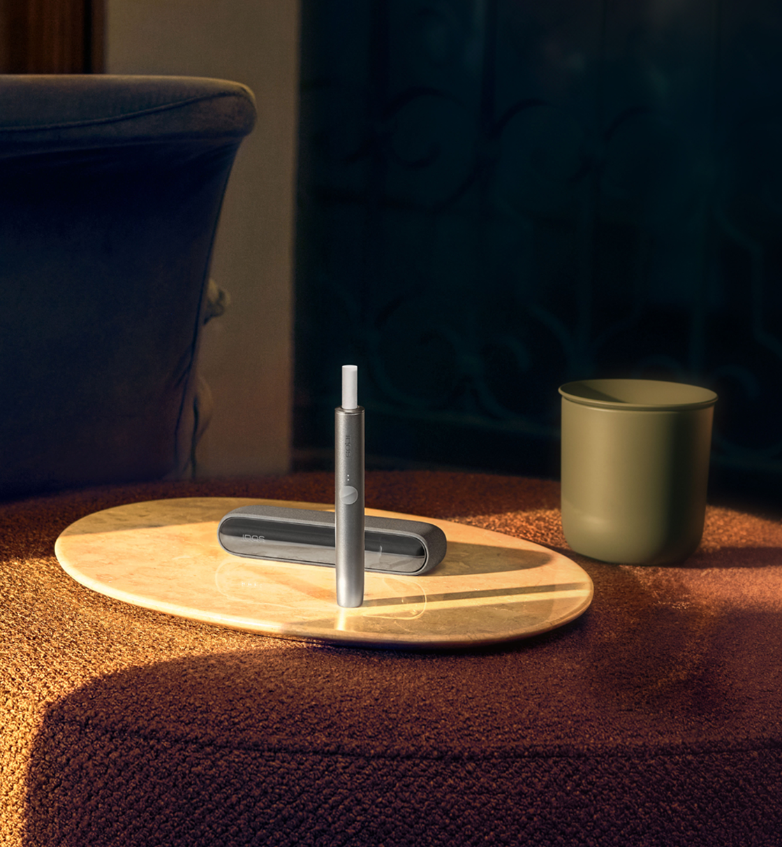Un appareil IQOS ILUMA debout avec un bâtonnet TEREA à l’intérieur et un plateau IQOS dans une pièce. 