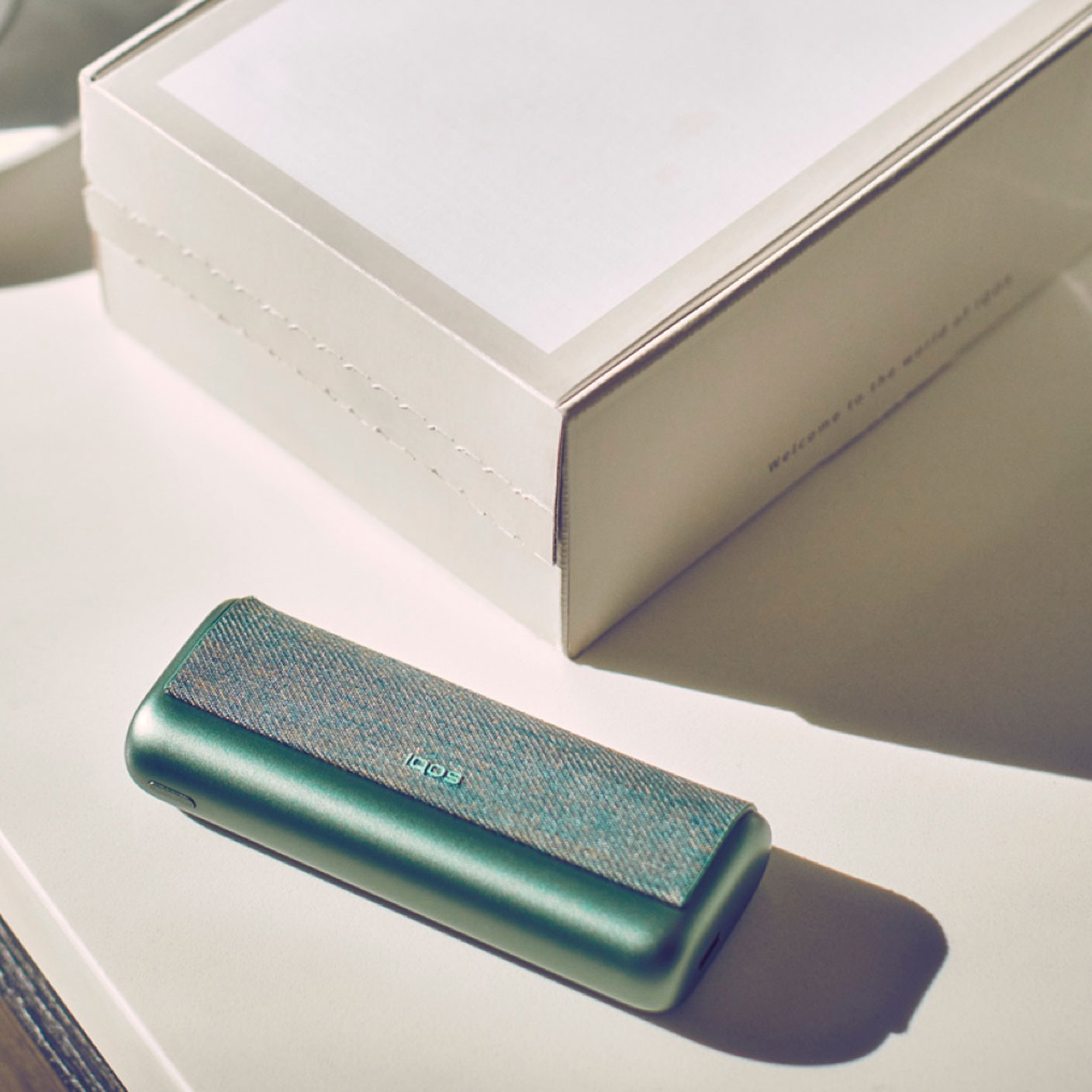 A jade green IQOS ILUMA PRIME Pocket Charger and an IQOS box on a table.