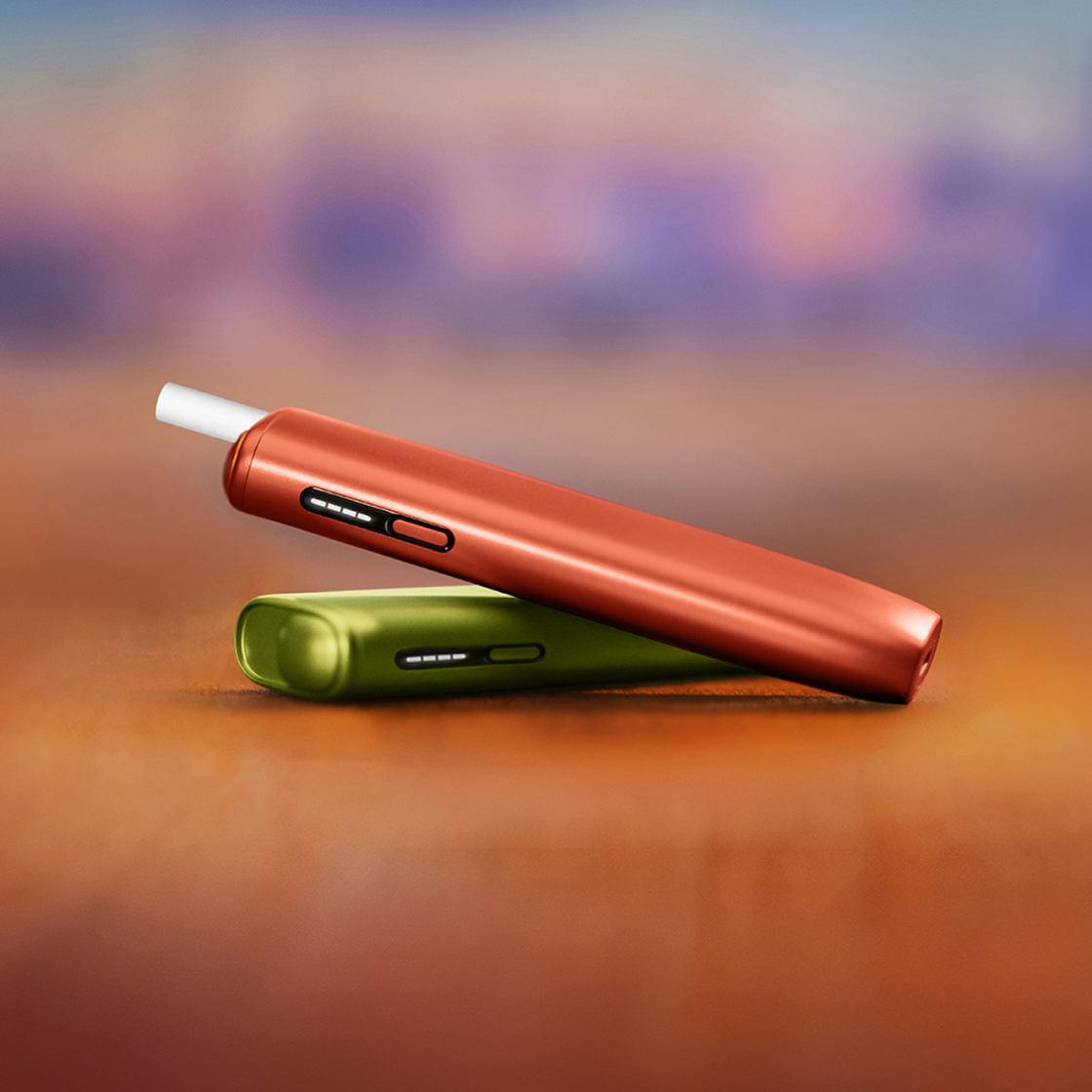 Vivid Terracotta IQOS ILUMA i ONE device with a TEREA stick inserted lying on top of an Aspen Green IQOS ILUMA i ONE in front of a gradient background. 