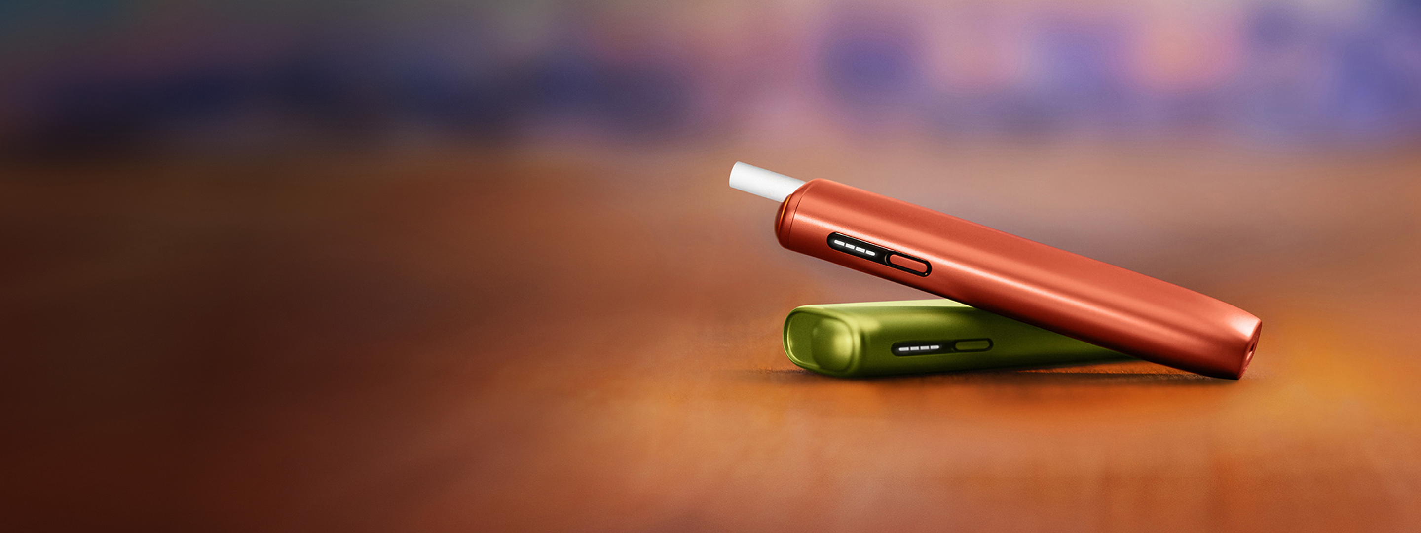 Vivid Terracota IQOS ILUMA i ONE device with a TEREA stick inserted lying on top of an Aspen Green IQOS ILUMA i ONE device on the floor.