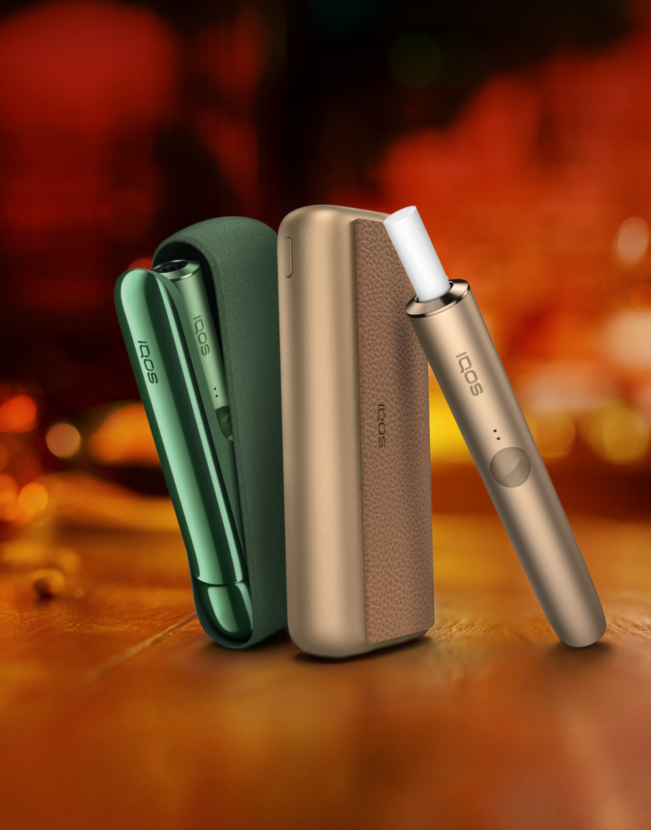 One green IQOS ILUMA device and one gold IQOS ILUMA device upright against a blurred orange/black background.